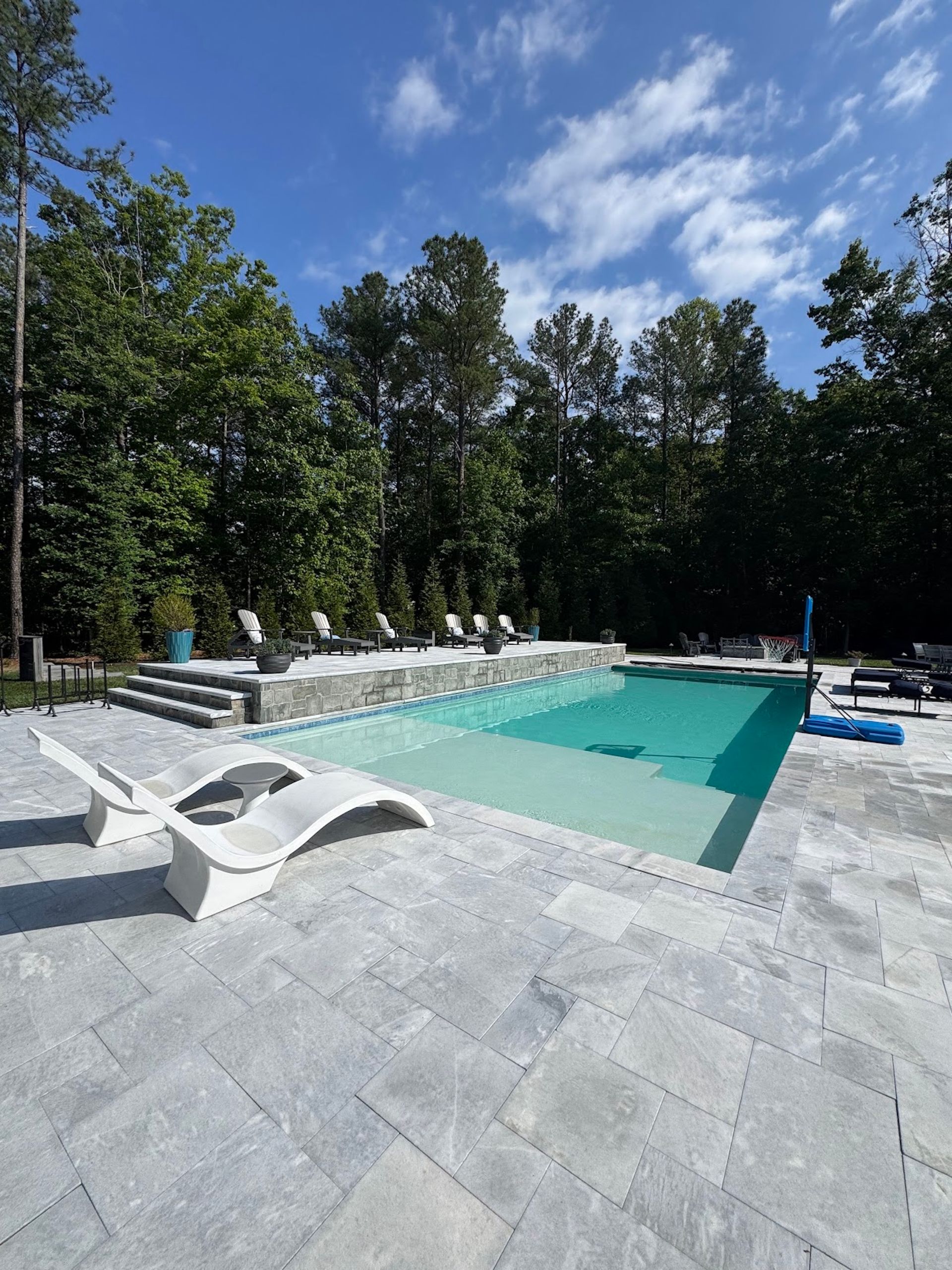 Outdoor swimming pool with turquoise water, white lounge chairs, and trees under a blue sky