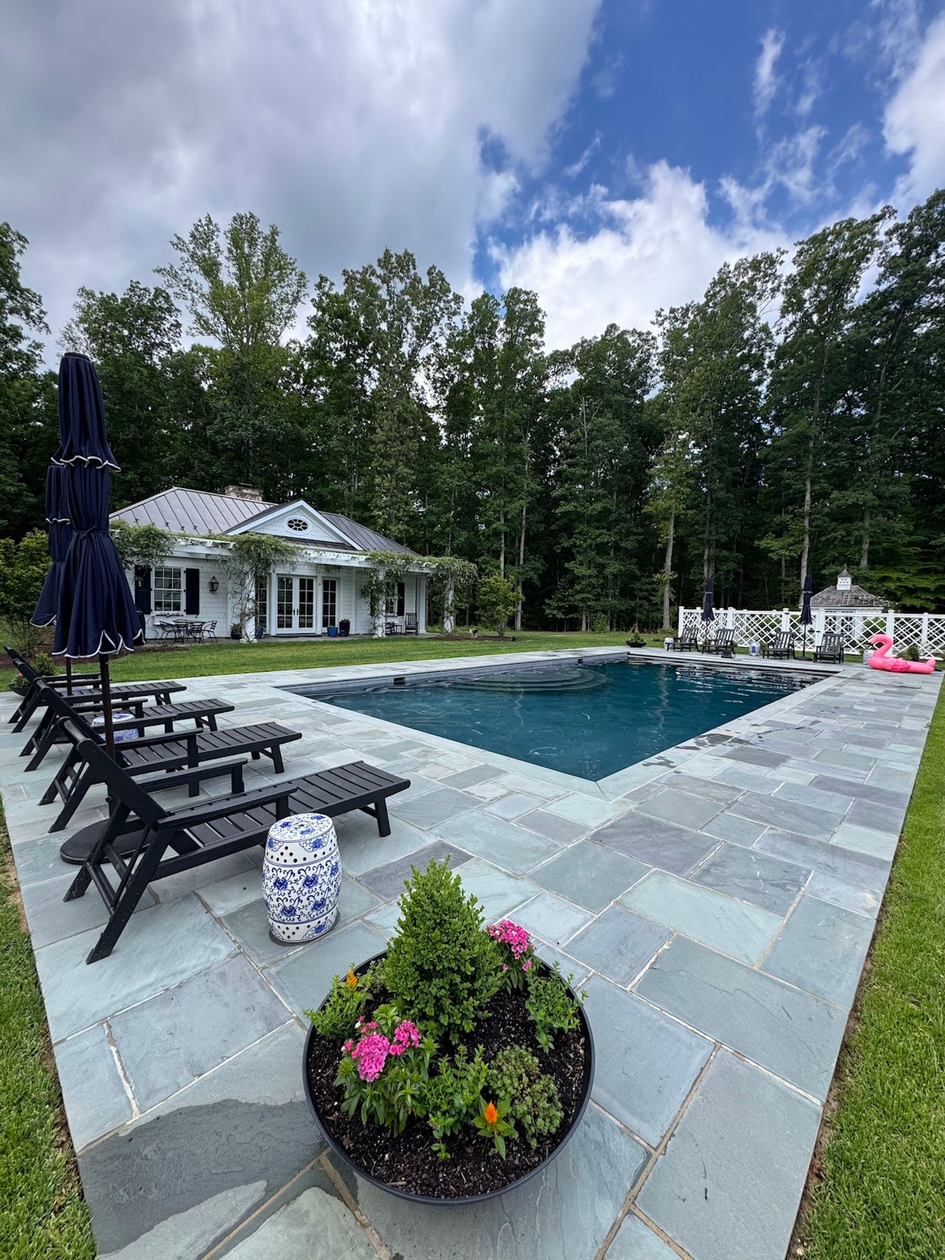 Backyard pool with black lounge chairs, tiled patio, white house, and trees under a cloudy sky