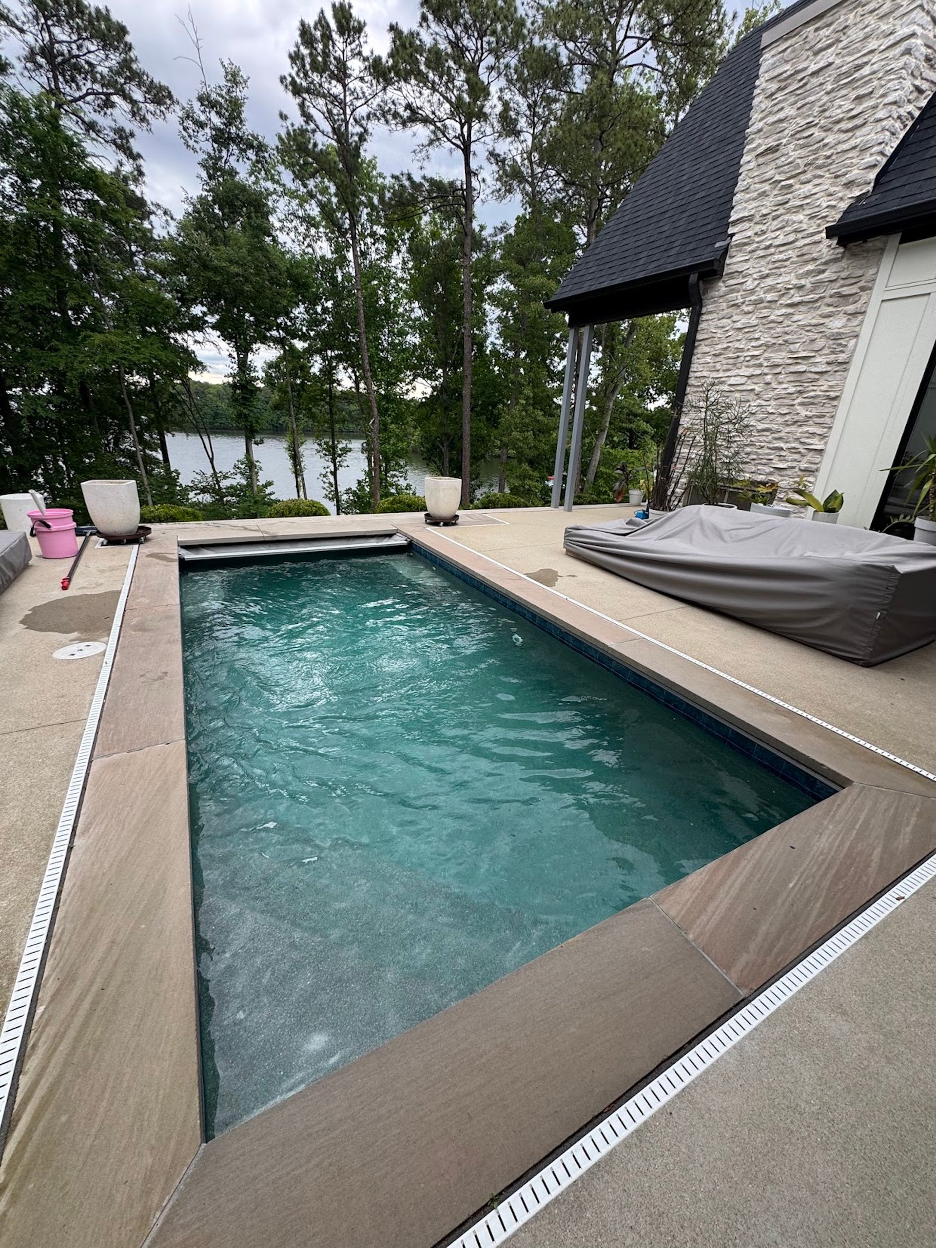 Backyard pool beside a stone house, with lounge chairs and trees in the background.