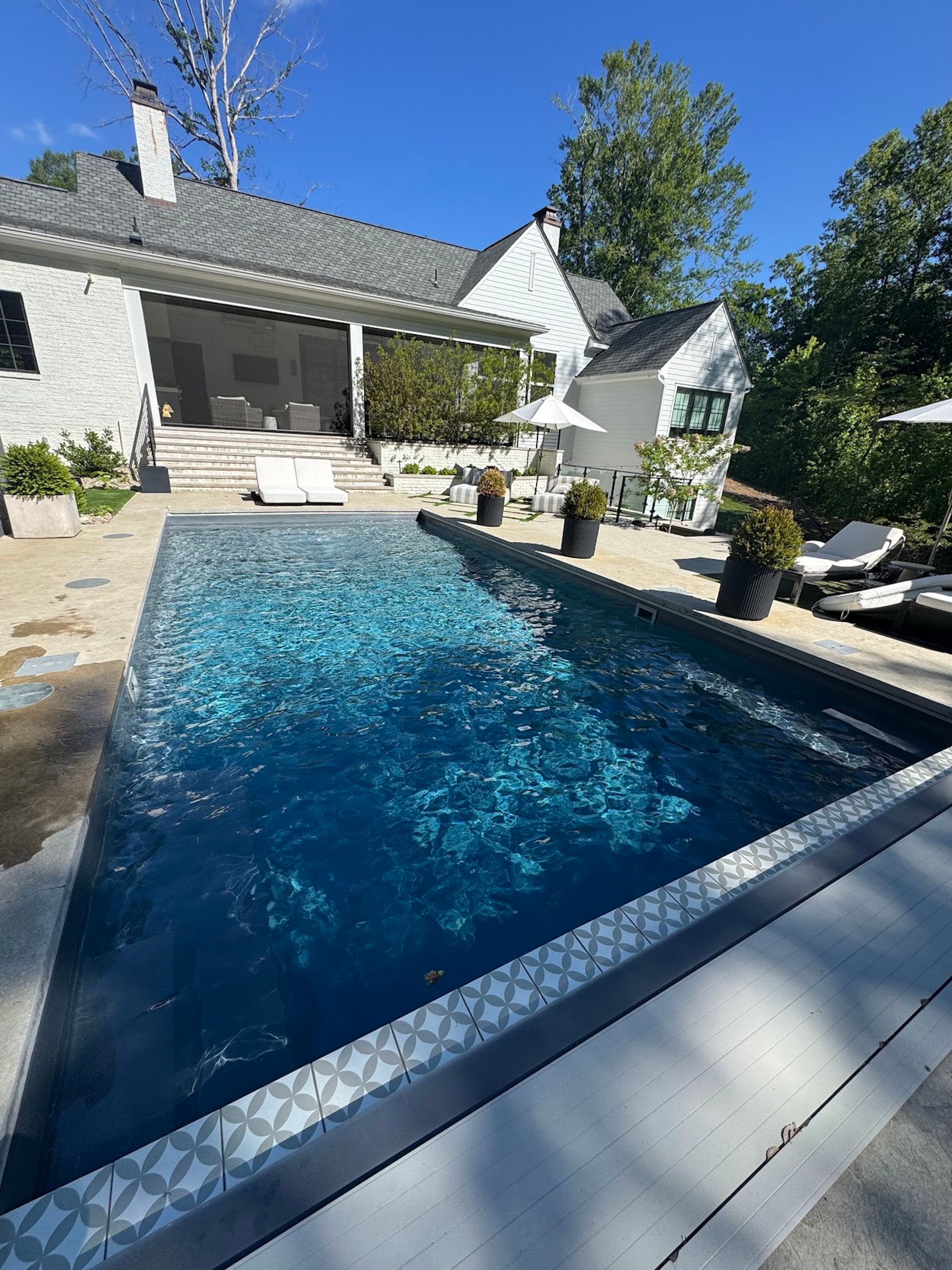 Backyard swimming pool beside a white house with patio seating and trees under a clear blue sky