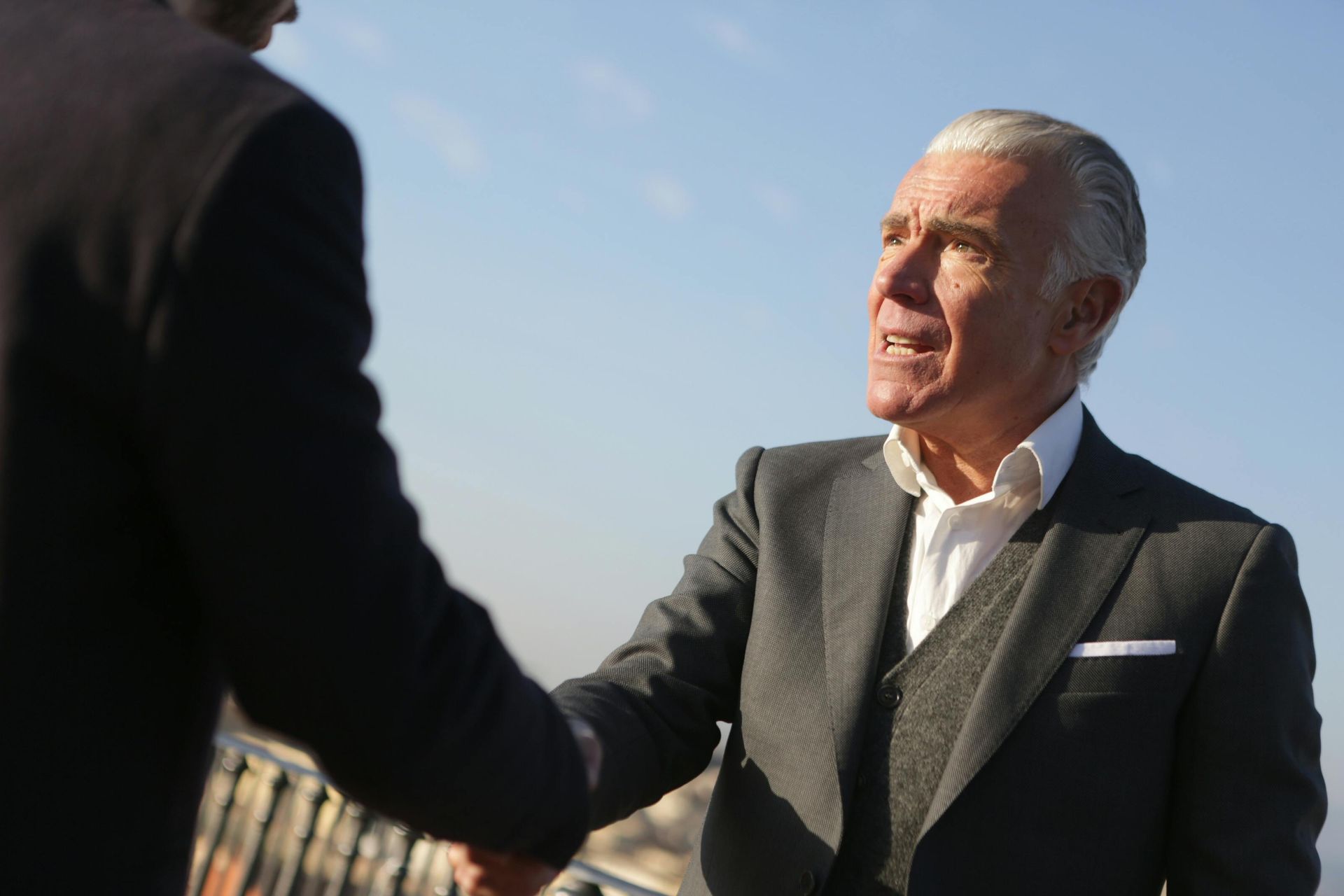 Two men in suits shaking hands outdoors with a blue sky background. One man has gray hair.