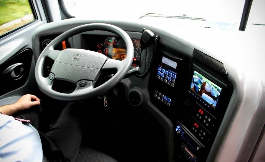 Cabine do motorista de ônibus com volante e painel de instrumentos com telas. O braço do motorista está visível.