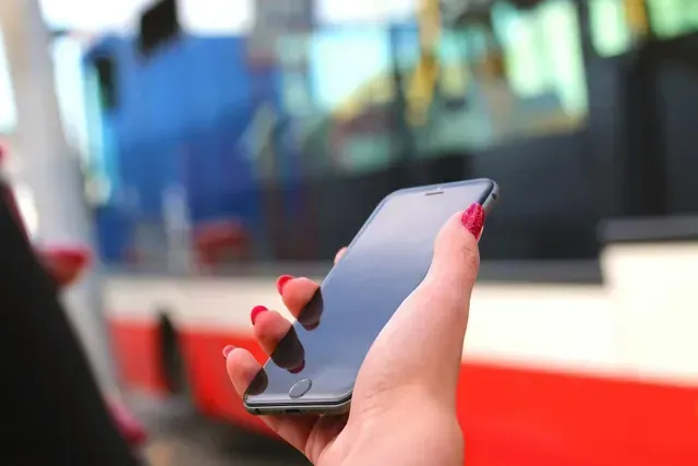 Mão segurando um smartphone em frente a um ônibus. Unhas esmaltadas de vermelho.