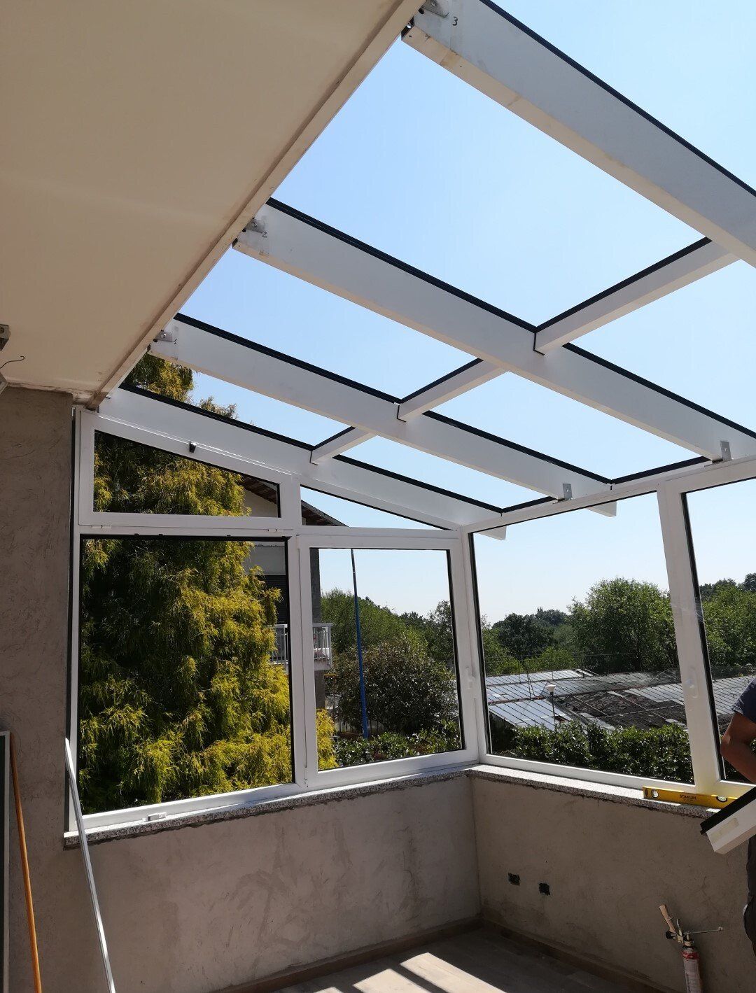 Patio con cornice bianca e tetto in vetro, con vista sugli alberi e sul cielo soleggiato.