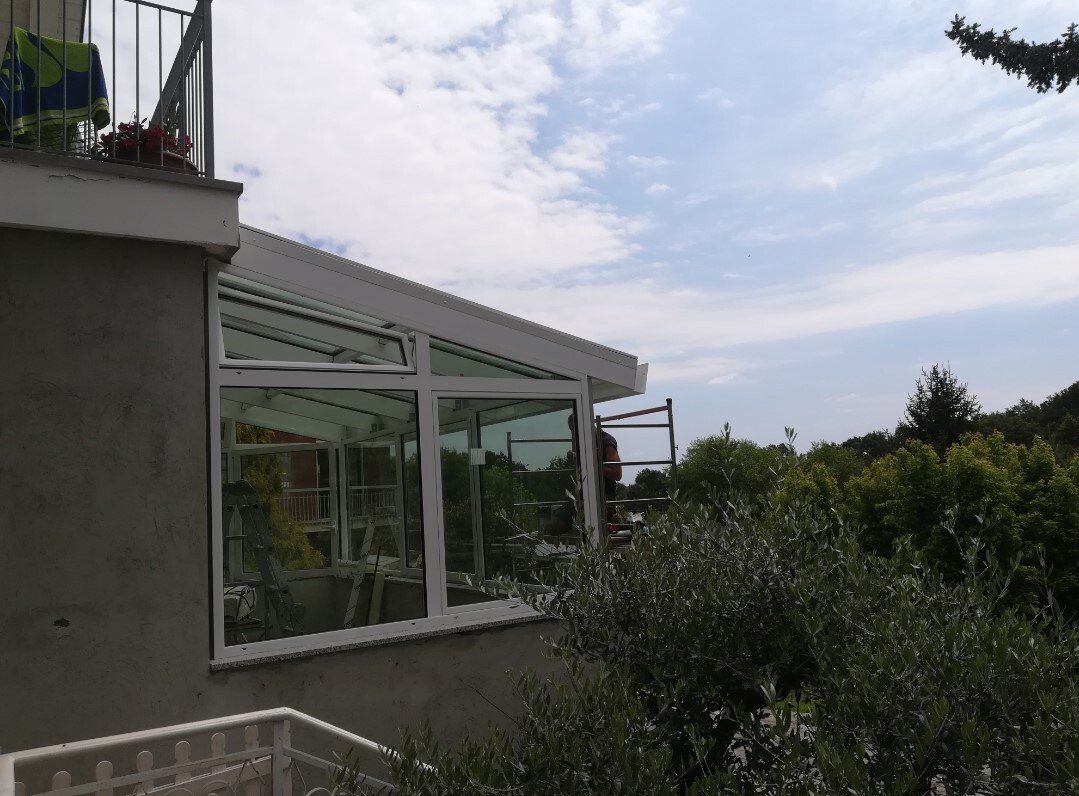 Veranda con pareti in vetro e cornice bianca, annessa all'esterno di un edificio. Con vista sugli alberi e sul cielo.