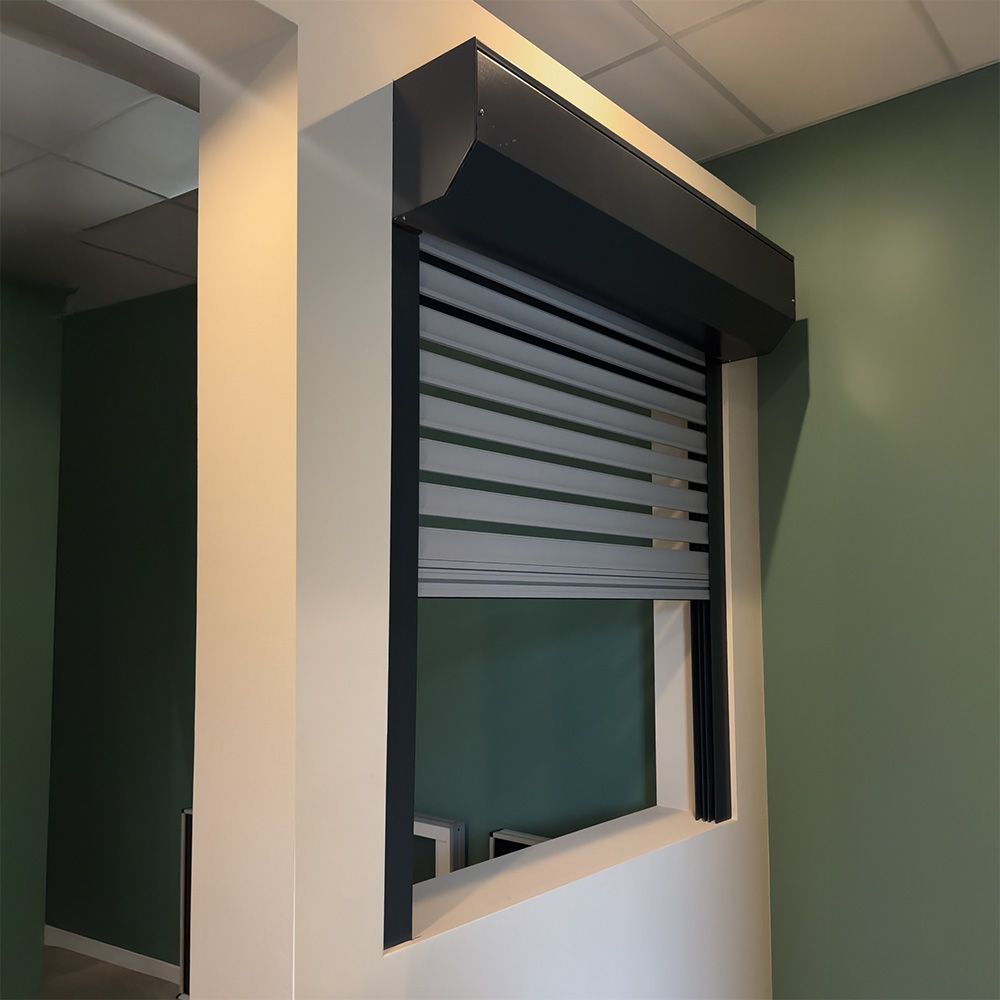 Una tapparella grigia copre parzialmente una finestra; installata su una parete bianca all'interno di uno spazio con pareti verdi.