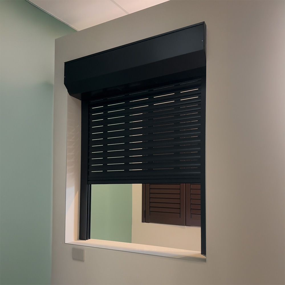 Una tapparella nera copre parzialmente una finestra in una stanza con pareti verde chiaro e beige.