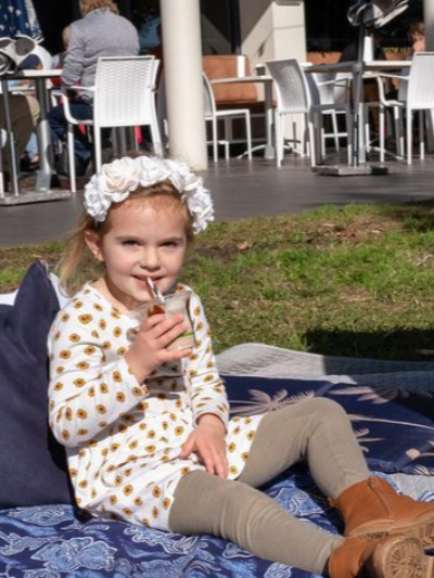 A young child wearing a floral crown and patterned dress sits on a picnic blanket drinking from a cup, outdoors. — Chop ‘n Chill Raby Bay in Cleveland, QLD