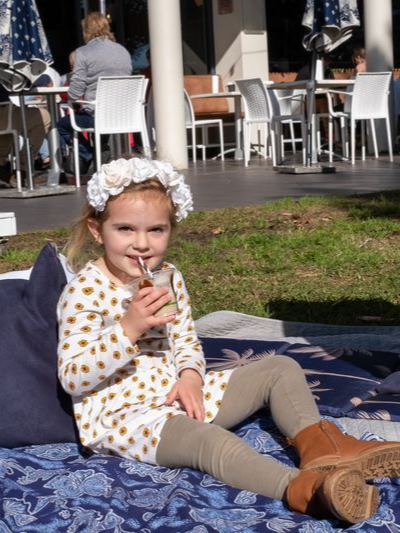 A young child wearing a floral crown and patterned dress sits on a picnic blanket drinking from a cup, outdoors. — Chop ‘n Chill Raby Bay in Cleveland, QLD