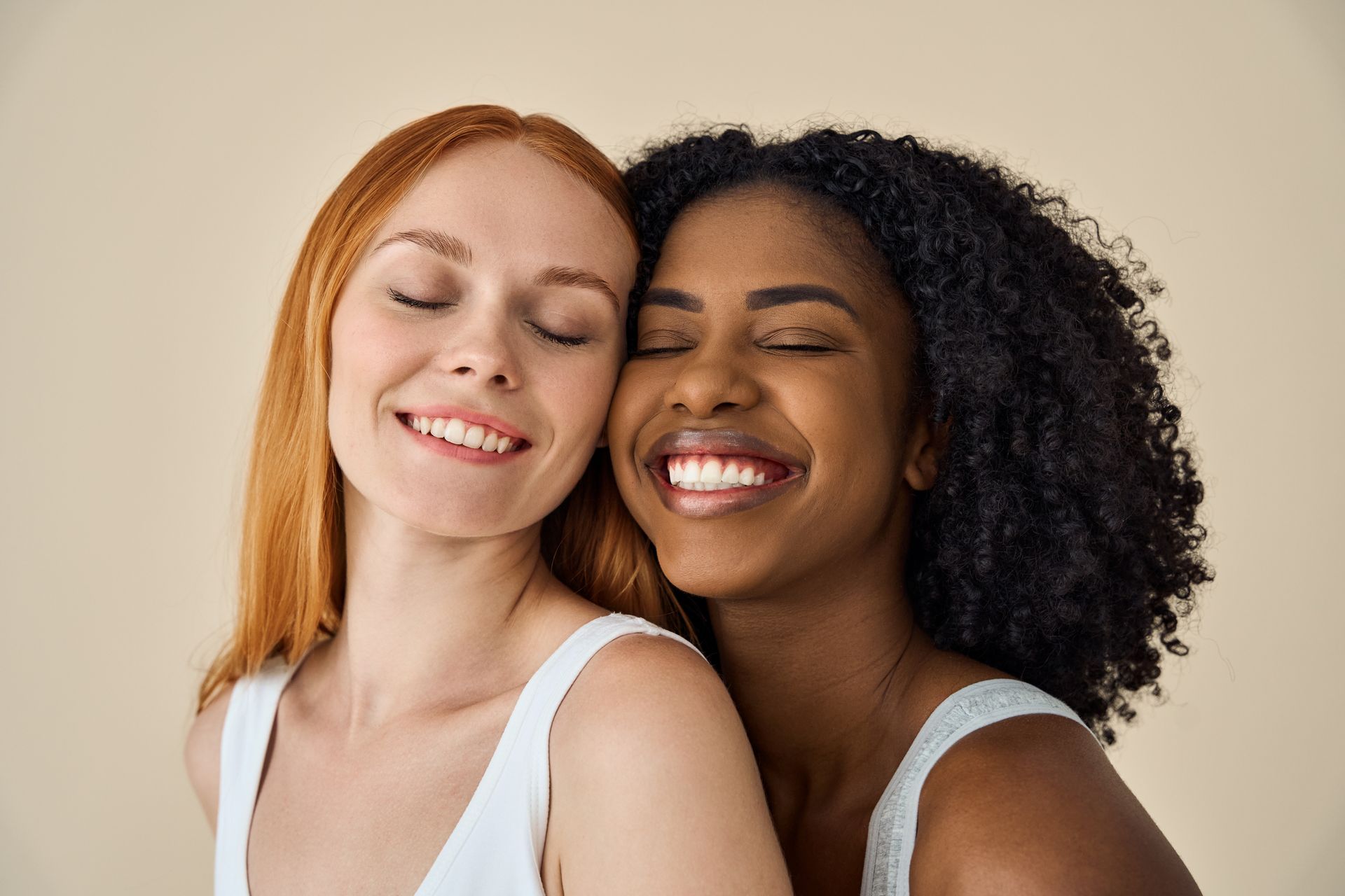 Two women smiling with eyes closed, heads touching. One has red hair, the other black. Beige background.