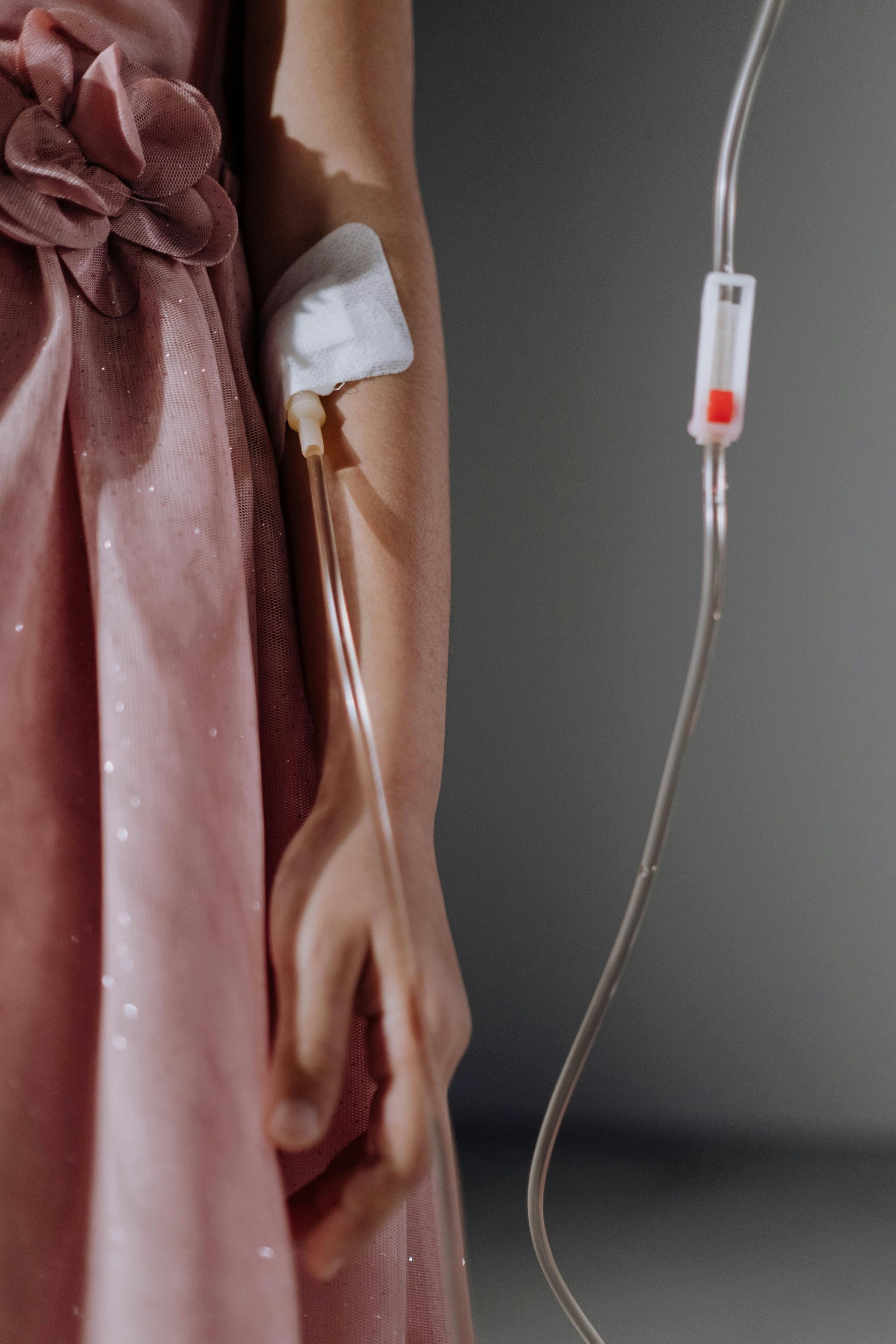 Person's arm with IV drip; wearing a pink dress. Cotton bandage on arm. Clear plastic tubing visible.