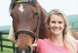 Woman smiling beside a brown horse in a sunny outdoor setting