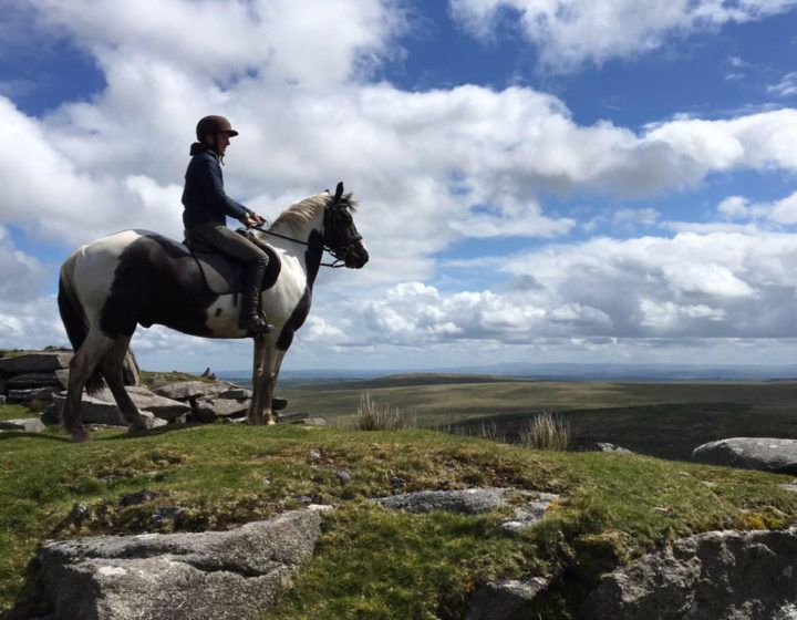 Person riding a black-and-white horse on a rocky cliff overlooking a wide landscape under a cloudy sky