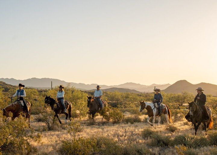 Five riders on horseback in a desert at sunset, with mountains in the background.