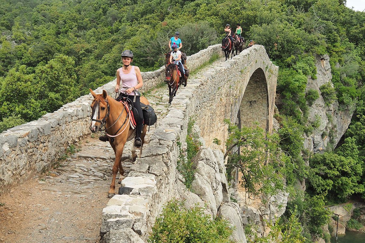 People horseback riding along a narrow stone bridge over a green valley