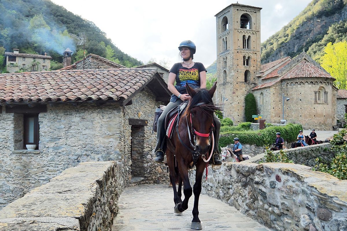 Person riding a horse over a stone bridge in a historic village with a church and mountains behind