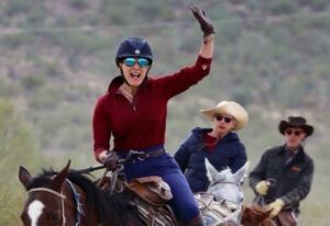 Three riders on horses, front rider in red raises a hand while smiling outdoors