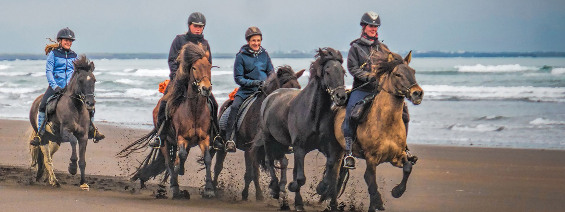 Four riders on horseback on a beach, with ocean waves in the background.