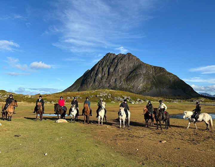Horse riders lined up on a grassy plain before a large rocky mountain under a blue sky.