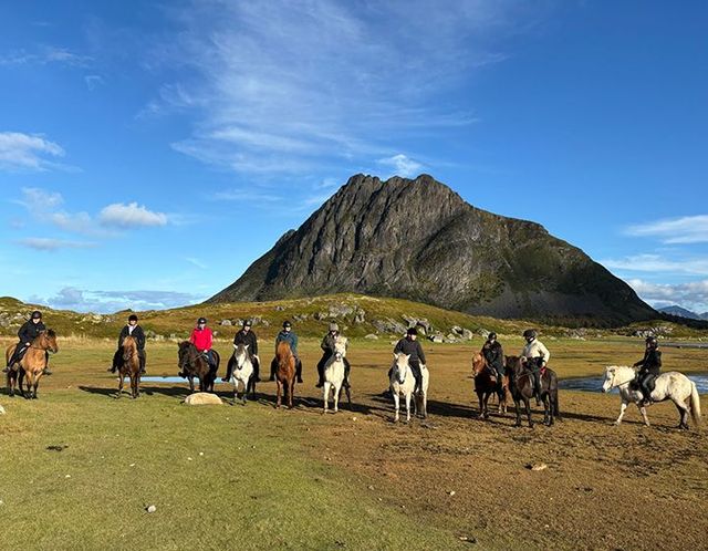 Riders on horseback in a grassy field with a large rocky mountain under a blue sky