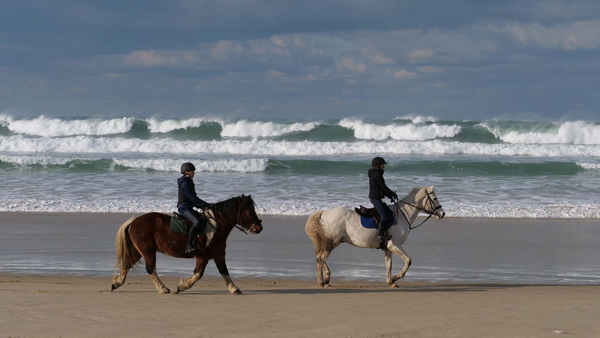 Two riders on horses walking along a sandy beach with waves breaking in the background
