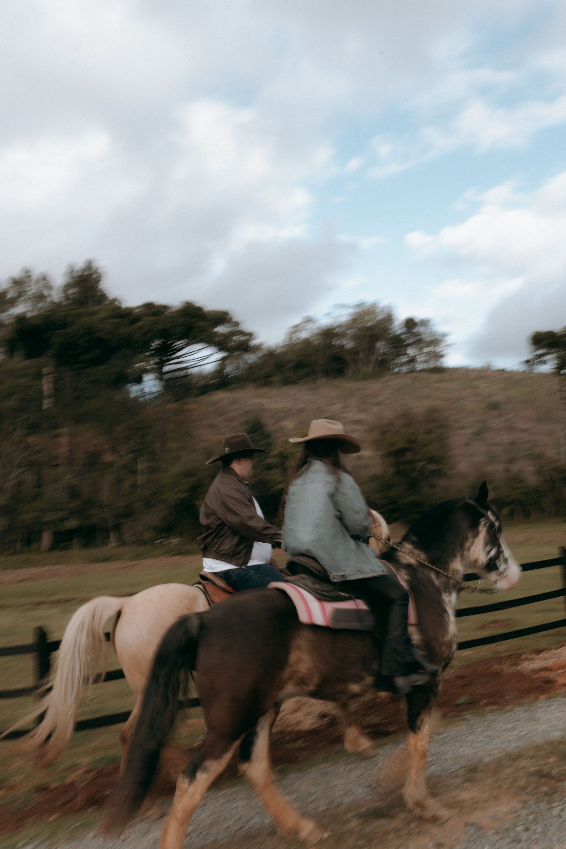 Two people riding horses on a dirt path beside a fenced field under a cloudy sky