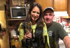 Two smiling people in a kitchen, holding wine glasses behind several wine bottles.