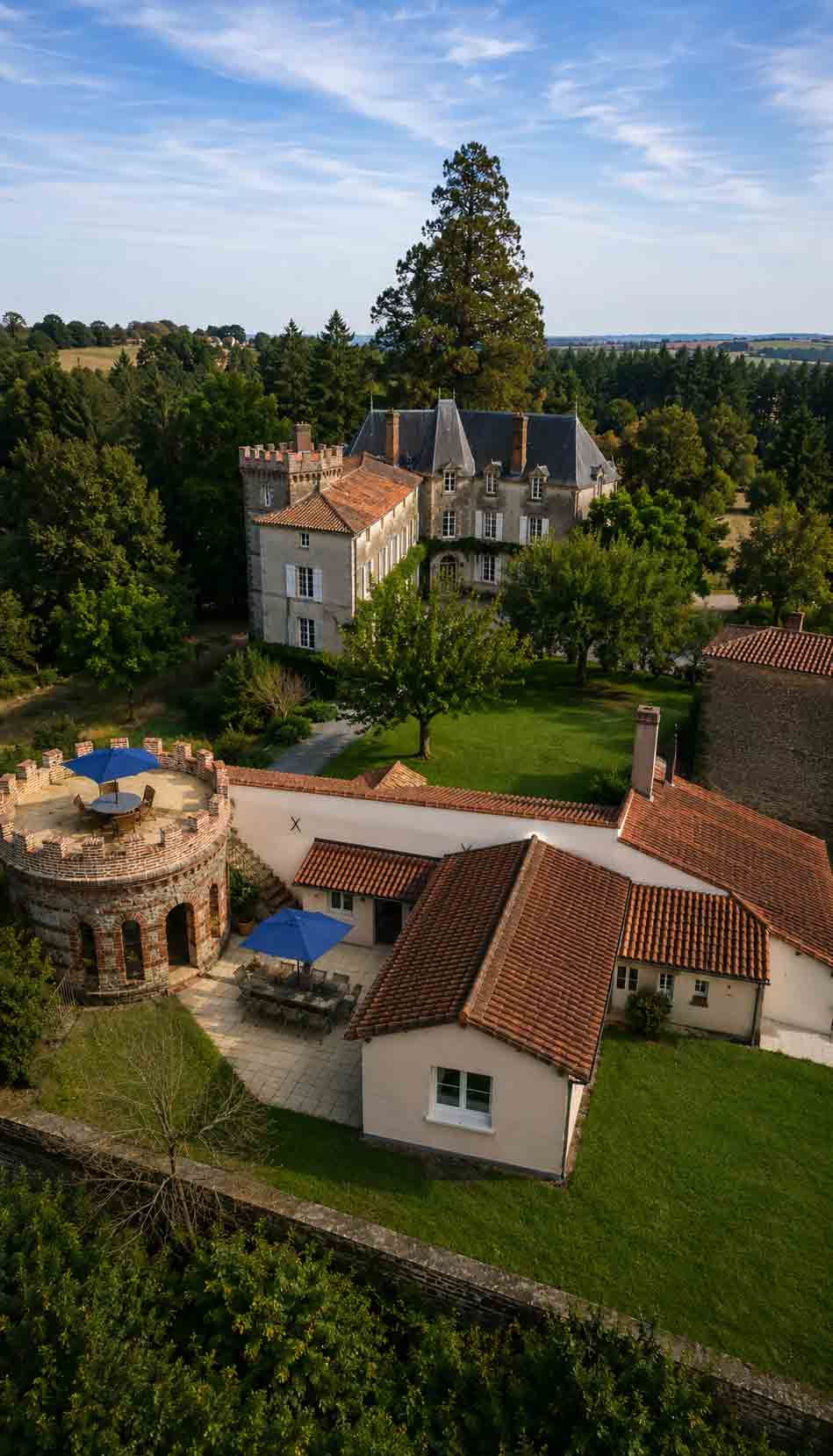 A large stone chateau with red tile roofs sits in a green, tree-filled landscape.