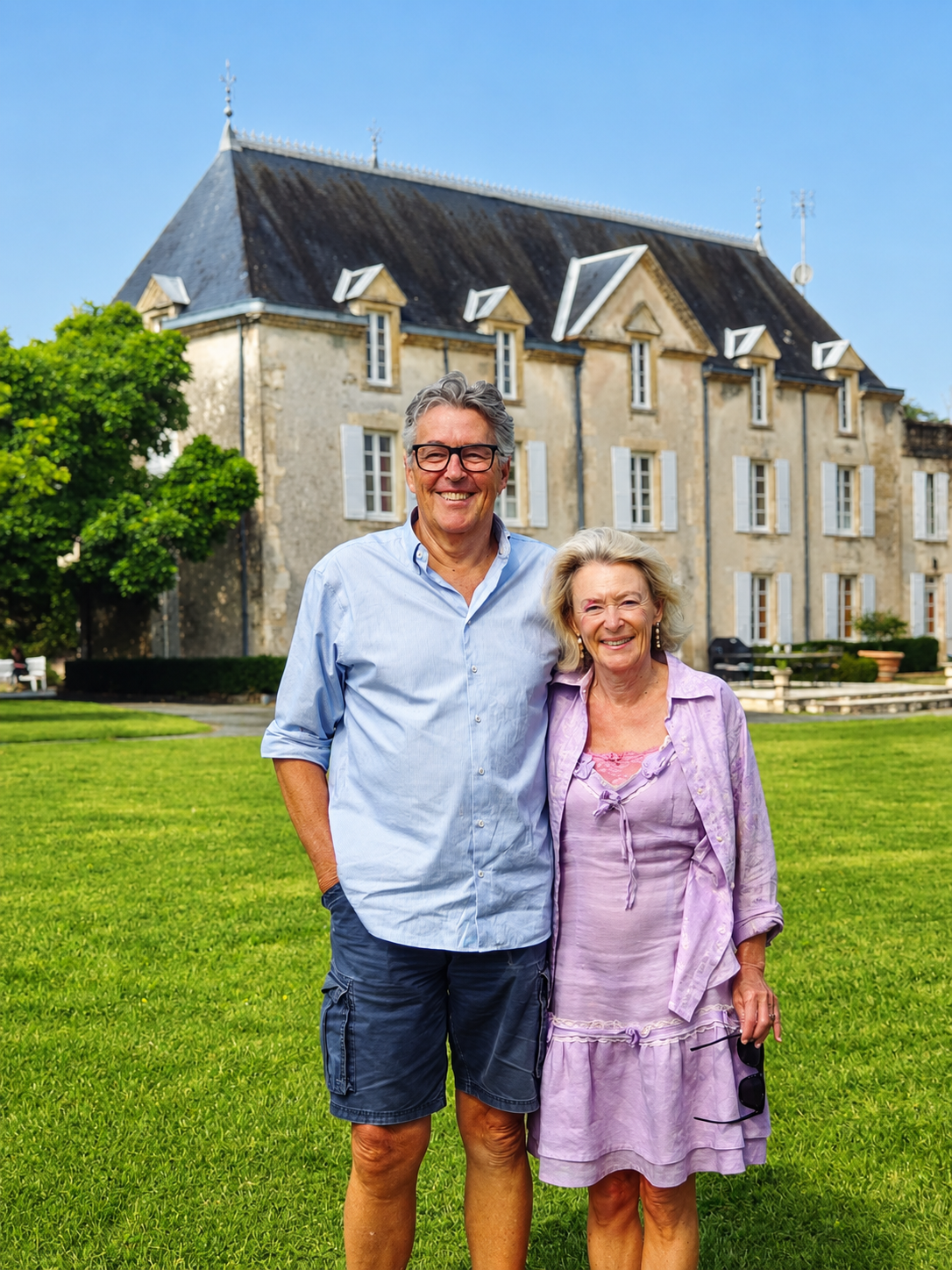 Couple posing in front of a large chateau. The man is wearing a blue shirt and shorts. The woman is wearing a purple dress.