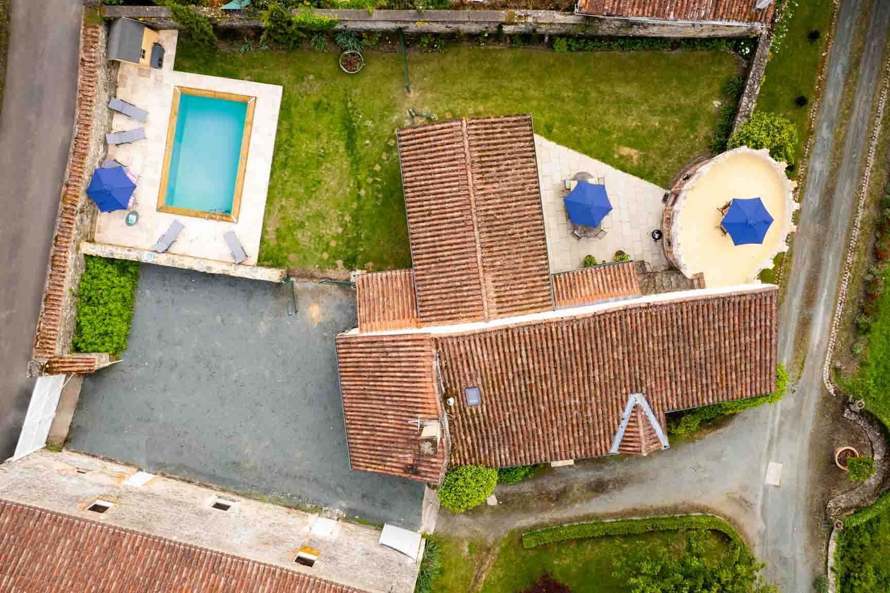 Aerial view of a house with red-tile roof, gray driveway, pool, and patio umbrellas in a fenced yard