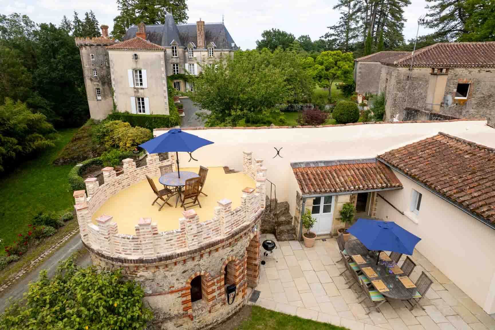 Stone château courtyard with round terrace, blue umbrellas, and outdoor dining tables amidst greenery