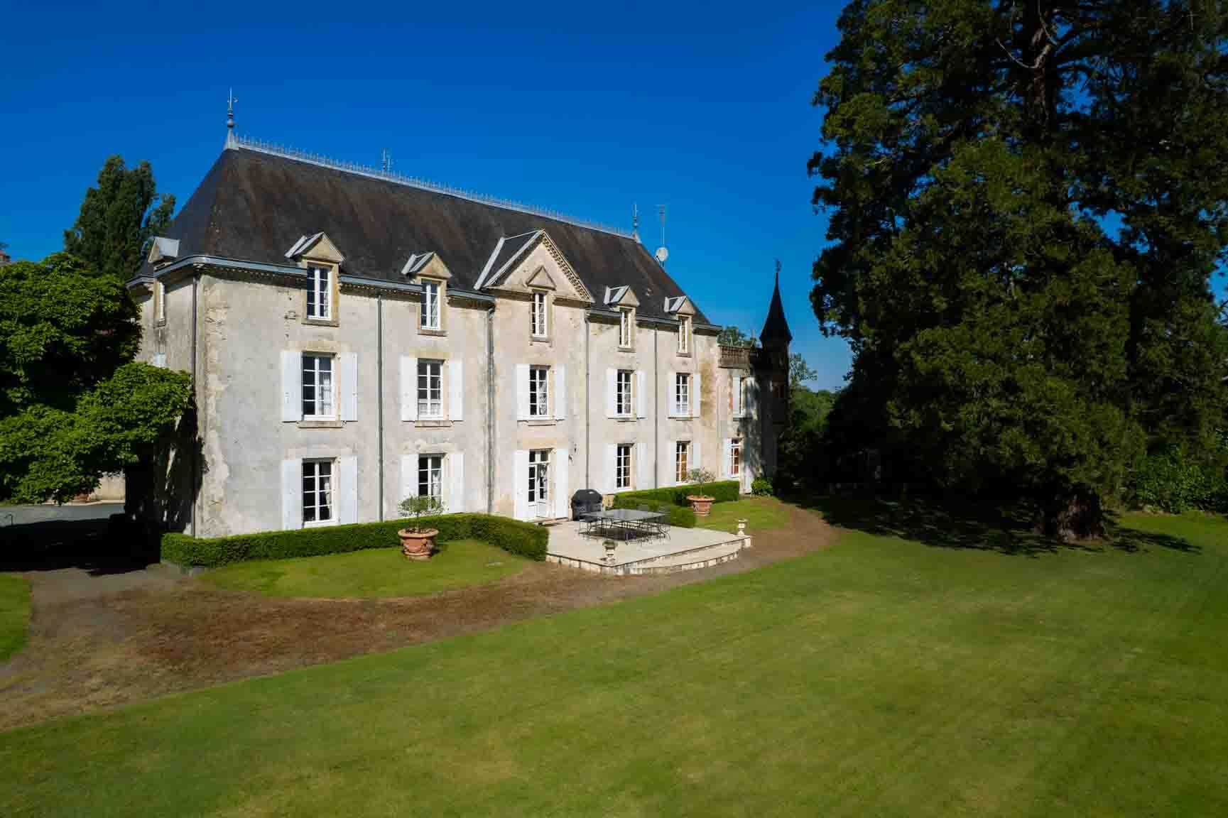 Large stone French chateau with a slate roof, set in a grassy landscape under a blue sky.