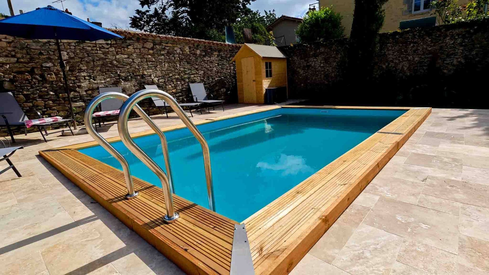 Backyard swimming pool with wooden deck, metal ladder, blue umbrella, and small shed behind a stone wall