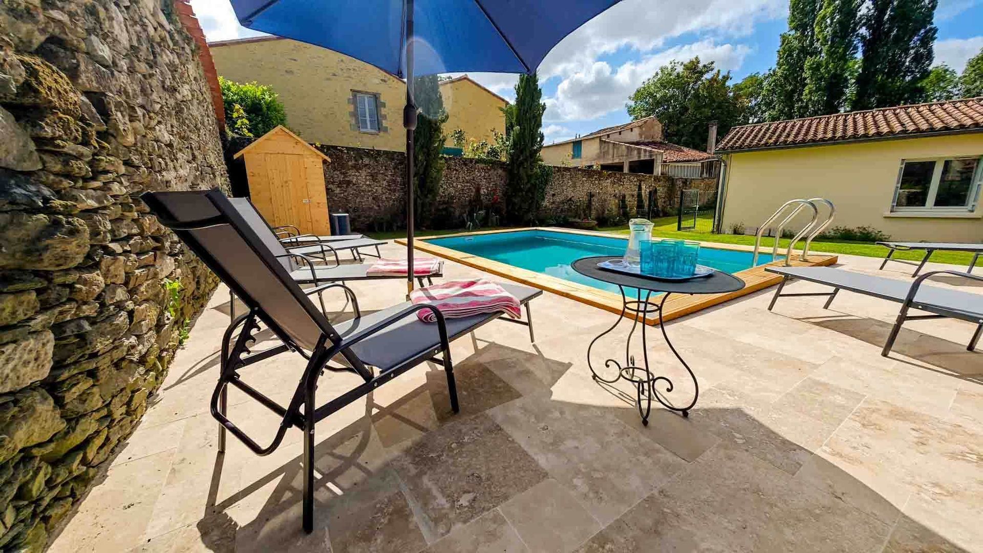 Patio with pool, lounge chair, table, and blue umbrella in a sunny garden courtyard