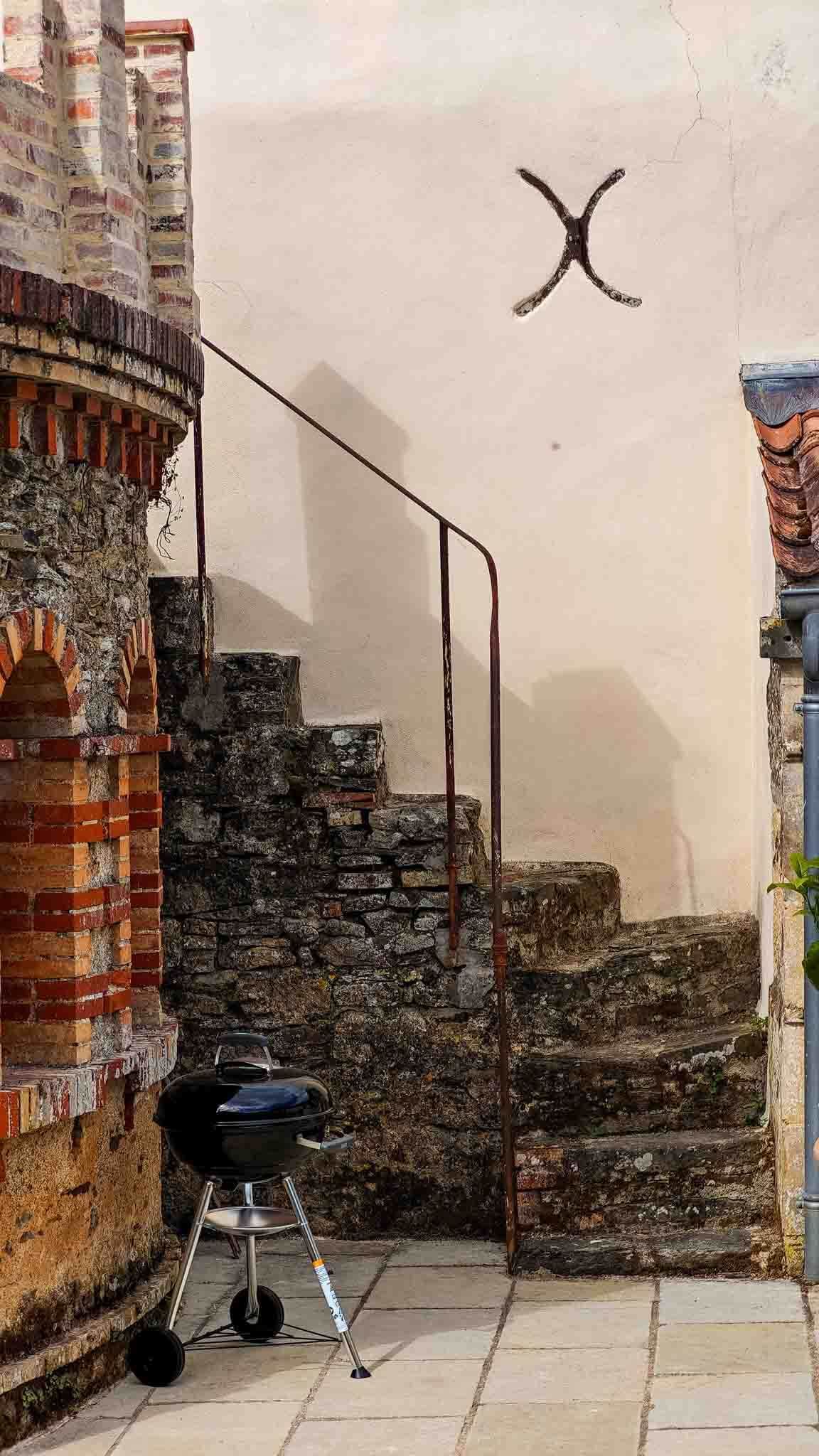 Stone staircase beside a brick building, with a grill at the bottom and a wall-mounted X-shaped mark above.