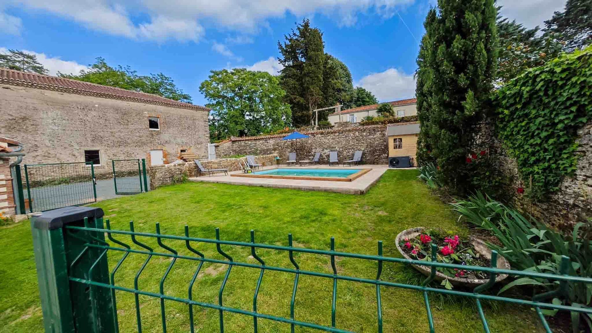 Backyard with green lawn, swimming pool, stone wall, and flower bed under a blue sky
