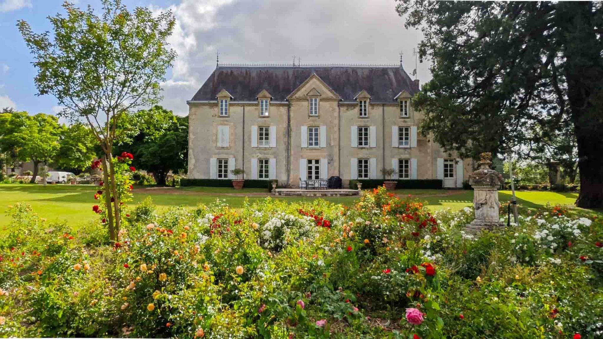 Elegant stone mansion behind a colorful flower garden and manicured lawn, framed by tall trees.