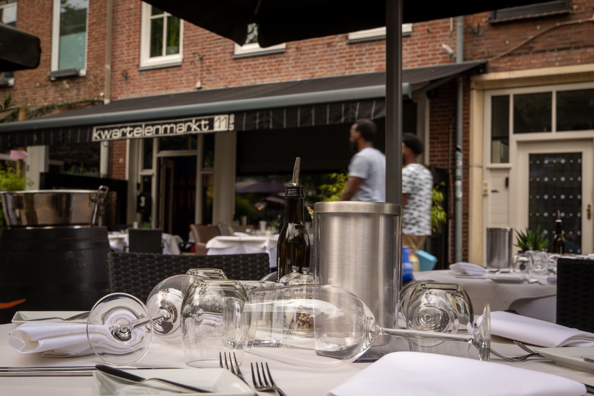 Restaurant in de openlucht, tafel met glaswerk, twee mensen op de achtergrond, voorgevel van het gebouw.