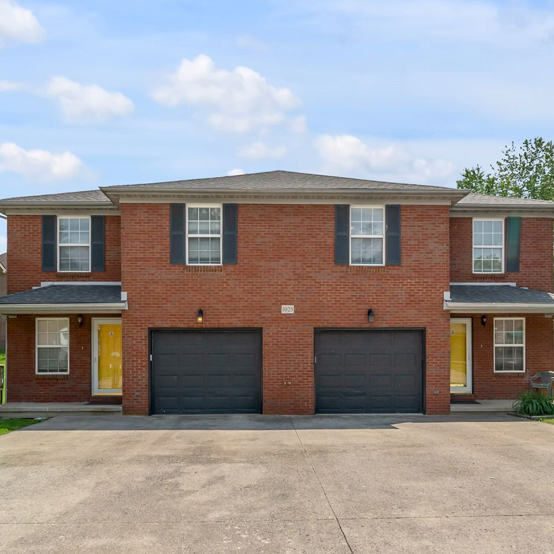 Two-story red brick duplex with black garages and a paved driveway under a partly cloudy sky