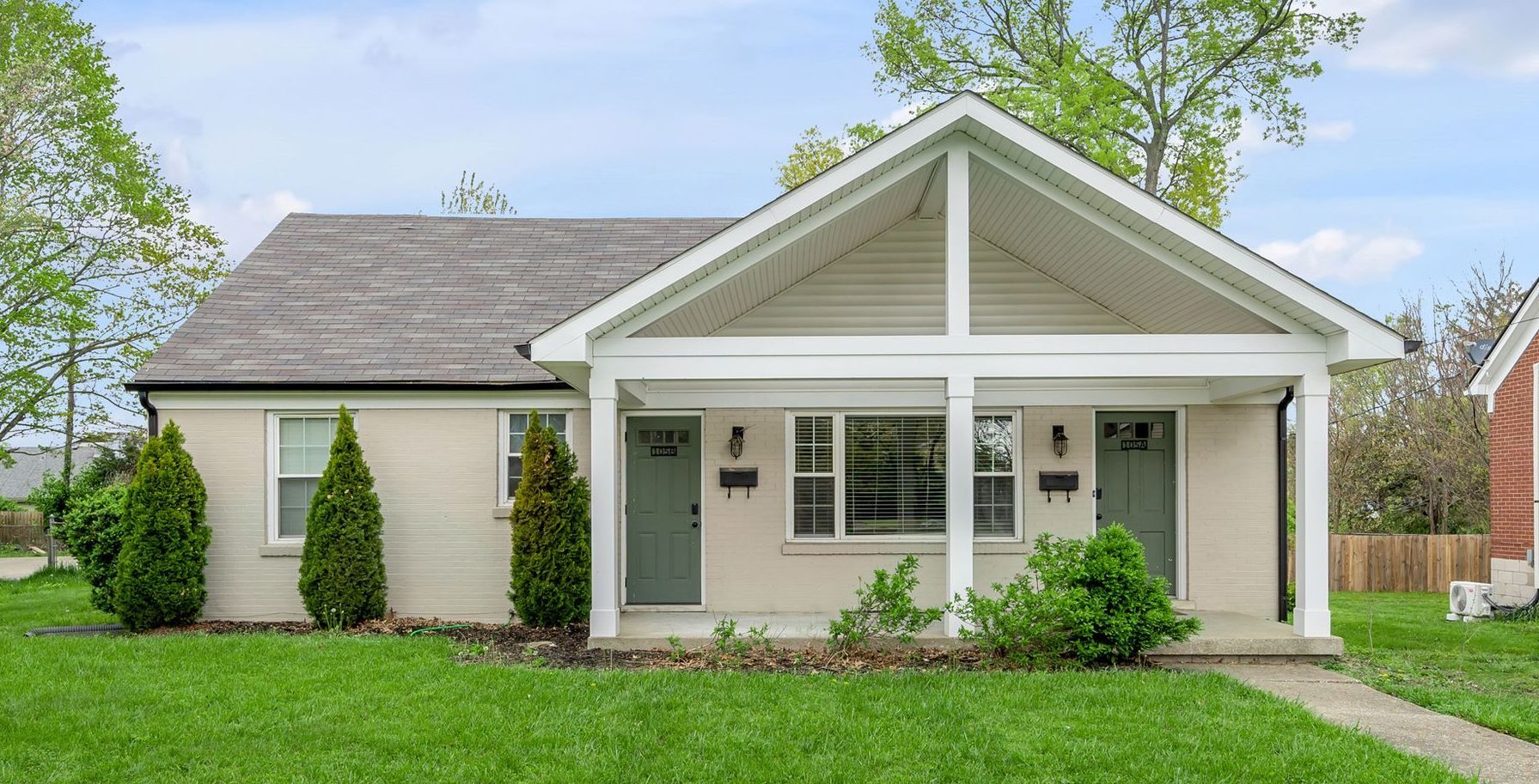 A light-colored brick duplex with a covered porch and green doors, set on a green lawn.