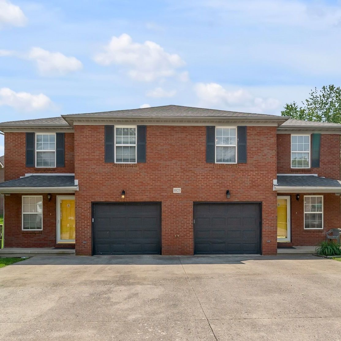Two-story red brick duplex with black garages and a paved driveway under a partly cloudy sky