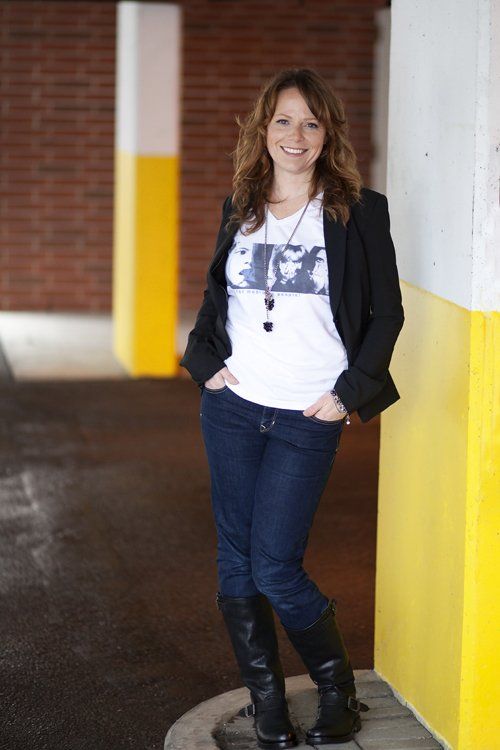 Woman standing in a hallway, leaning against a wall, wearing a black jacket, white shirt, jeans, and boots