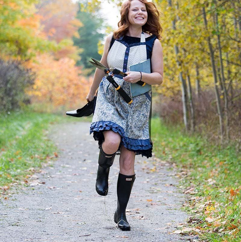 Woman in patterned dress walking with a camera on a leafy autumn path, smiling.