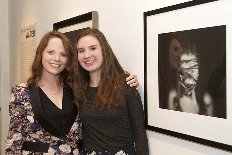 Two women smiling beside a framed black-and-white portrait in a gallery.