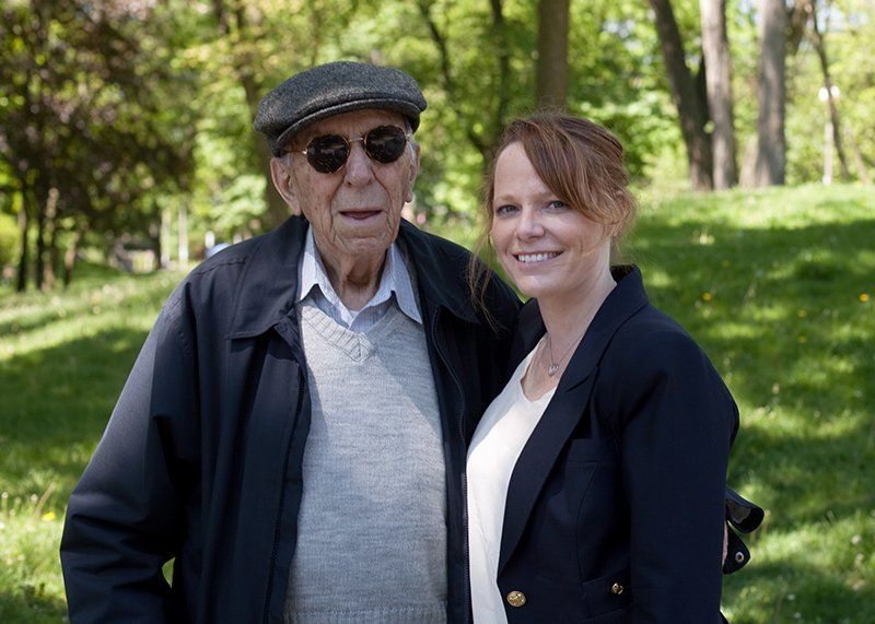 Two people standing together in a sunlit park, one wearing a cap and sunglasses, the other in a dark blazer.
