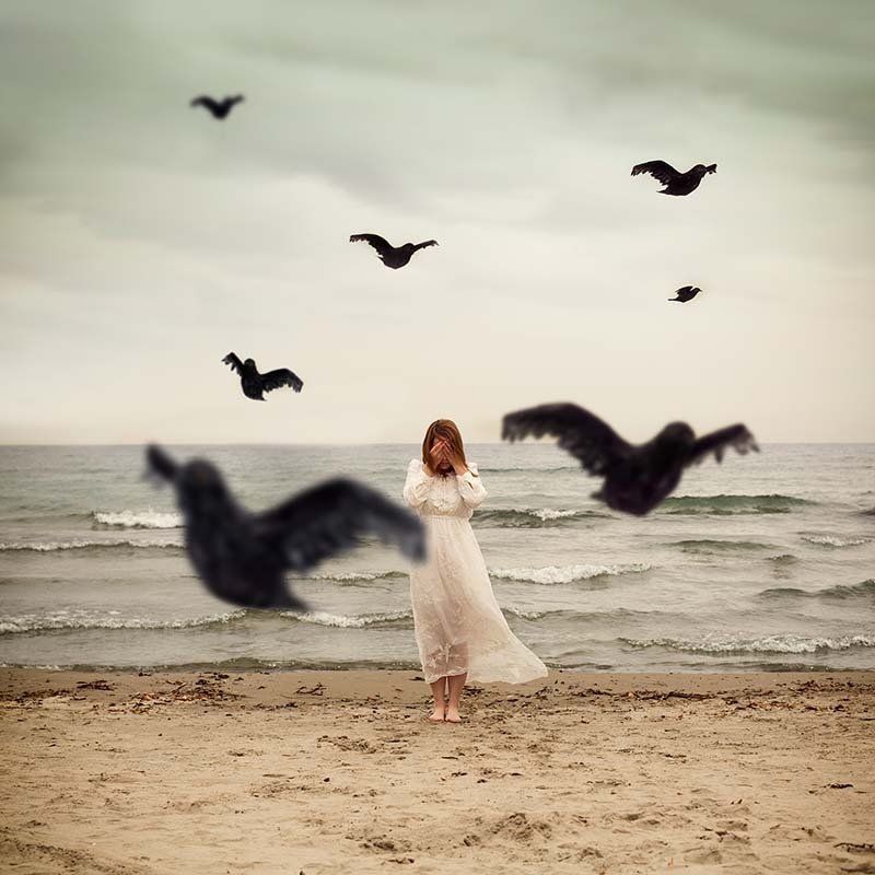 Woman in a flowing white dress stands on a sandy beach as birds fly overhead near the ocean.