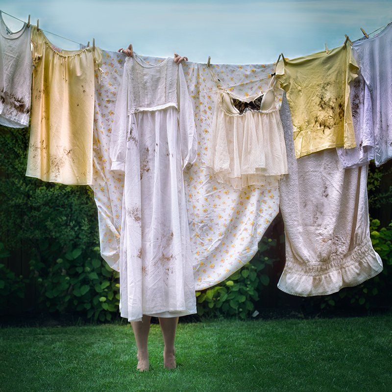 Laundry hanging on a clothesline in a grassy yard, with shirts, dresses, and sheets in white and pale yellow