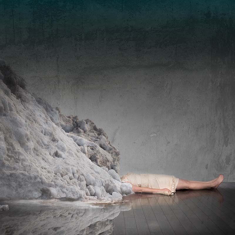 Person lying on a floor beside a large gray-white mound against a dark wall