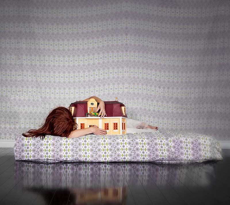 Dollhouse on a patterned bedspread with a doll lying beside it against a gray patterned backdrop
