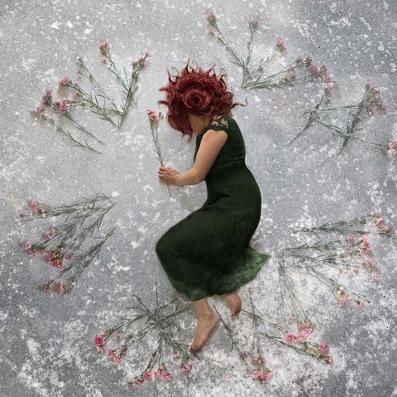 Woman in a dark green dress lying curled on a snowy surface, surrounded by scattered red flowers and branches.